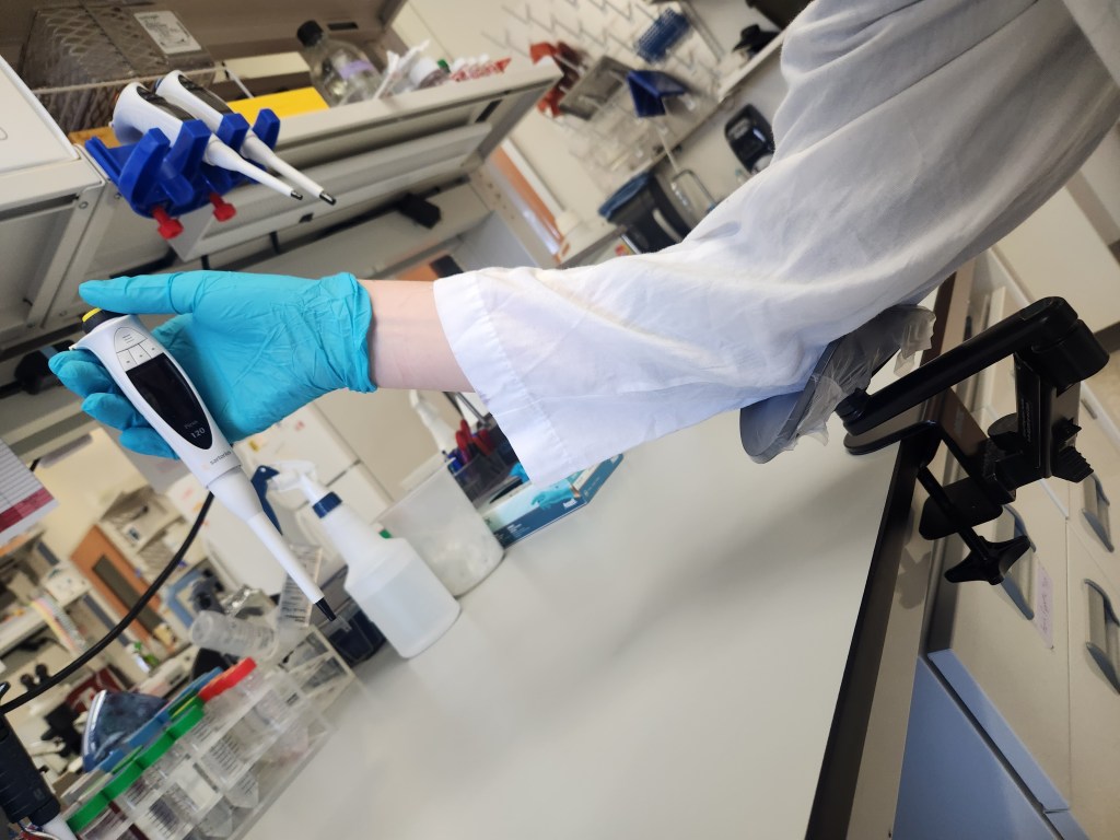 A scientist in a white lab coat and teal gloves holds a pipettor while resting their elbow on the arm rest, which is attached to the edge of the lab bench.