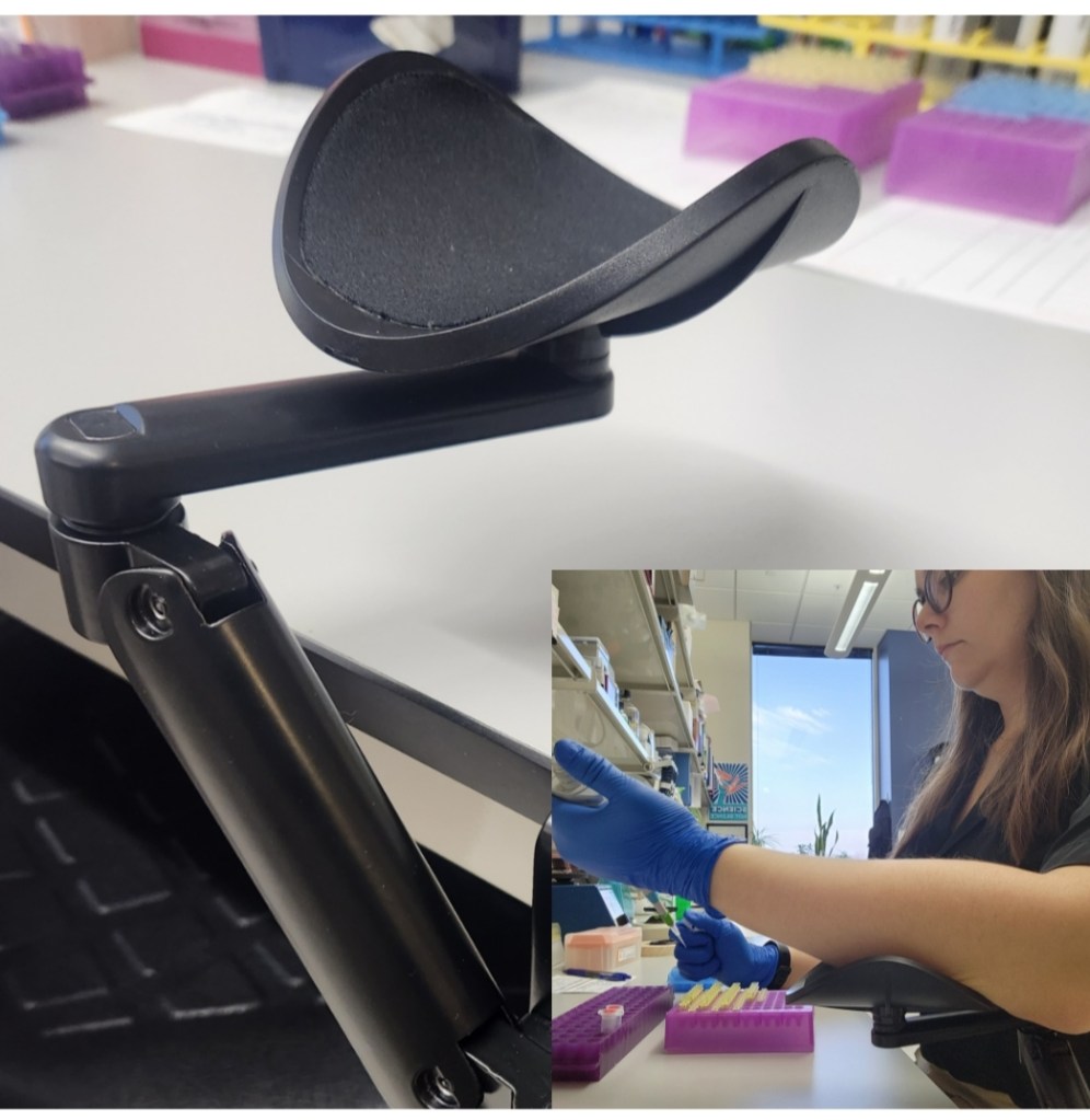 A black arm rest is attached the a grey lab bench. In the right hand corner, Kat demonstrates usage of the arm rest by placing her right forearm on it while she pipettes with a blue glove on.