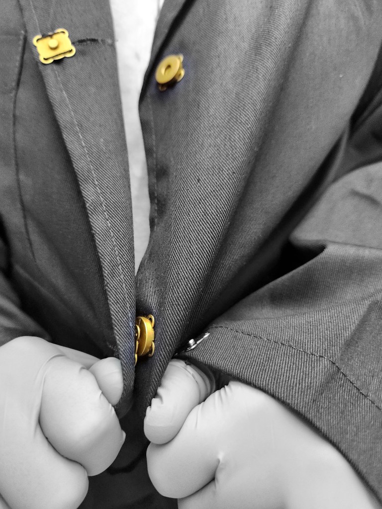 A zoomed in black and white photo of an individual’s gloved hands buttoning a lab coat. The lab coat contains magnetic buttons, which appear in gold