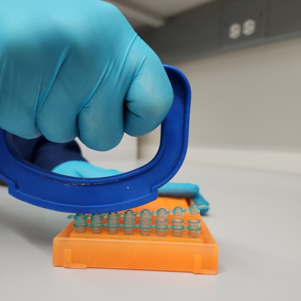 A teal gloved hand holds a bright blue half-moon shaped plastic tool (cap it tool) via a handle. The hand is pushing down on the blue tool, to attach a strip of green lids to green tubes via a groove on the Cap-it Tool opposite the handle. The green tubes/lids reside in a bright orange grid.