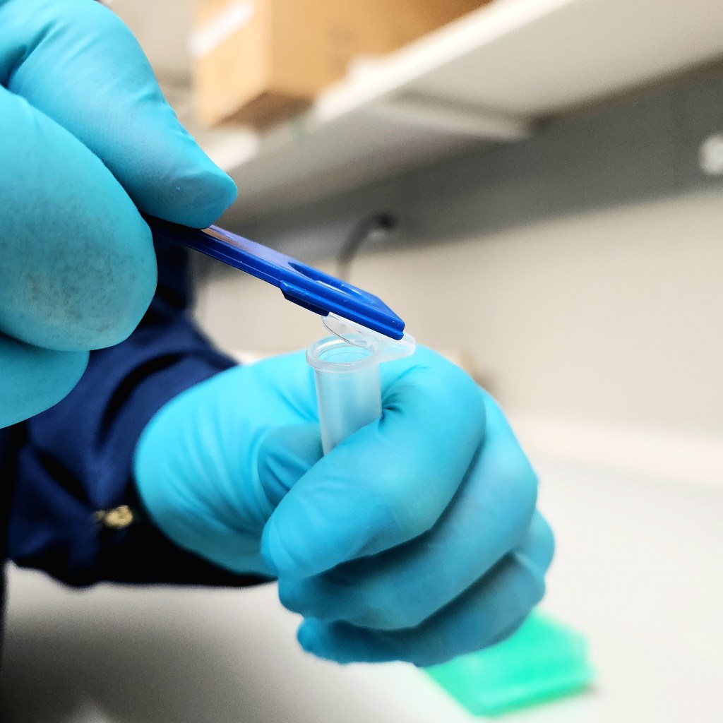A teal gloved hand uses a dark blue, plastic lever to open the Eppendorf tube.